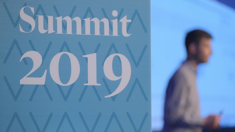 photo of a speaker on stage. The speaker is in the background and blurred. In there foreground is a sign reading "Summit 2019"