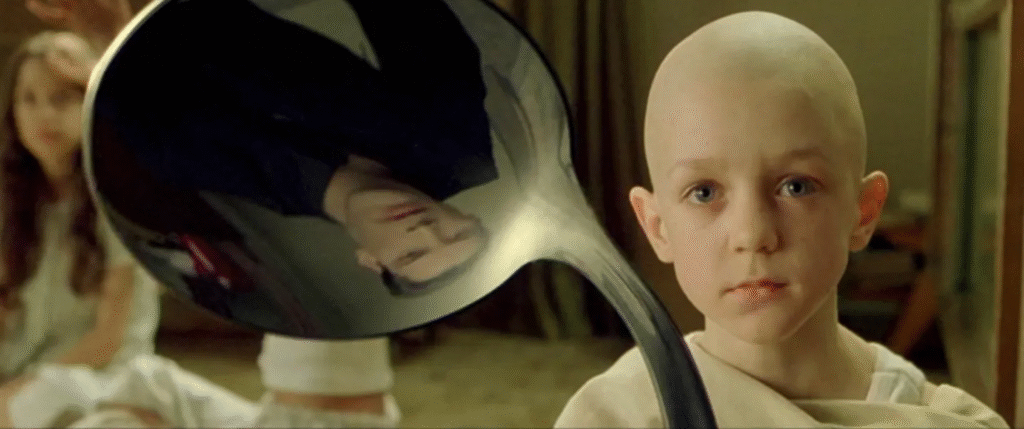 Bald child in a beige robe holding a bent spoon reflecting the image of a man in black, with a blurred figure in white seated in the background, from a scene in The Matrix.